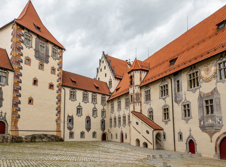 Das Bild zeigt den Innenhof des Hohen Schlosses in Füssen, der durch seine Illusionsmalerei an den Fassaden beeindruckt. Das Schlossgebäude hat rote Türen und ein Ziegeldach.