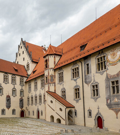 Das Bild zeigt den Innenhof des Hohen Schlosses in Füssen, der durch seine Illusionsmalerei an den Fassaden beeindruckt. Das Schlossgebäude hat rote Türen und ein Ziegeldach.