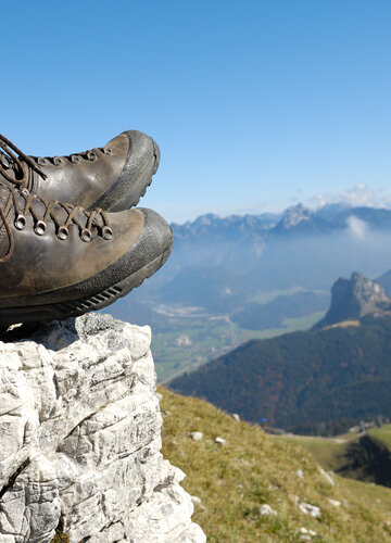Das Bild zeigt die ledernen Bergschuhe einer Person, die auf einem Berg sitzt. Weiter unten erkennt man Bergwiesen, Wälder und ein Tal. Dahinter erheben sich weitere Berge. 
