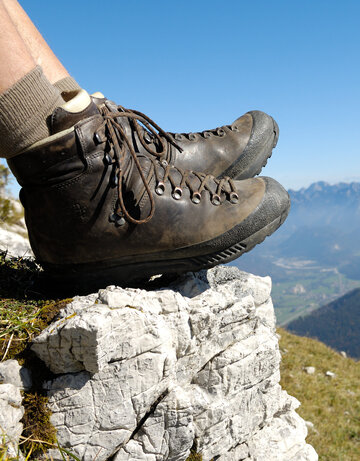 Das Bild zeigt die ledernen Bergschuhe einer Person, die auf einem Berg sitzt. Weiter unten erkennt man Bergwiesen, Wälder und ein Tal. Dahinter erheben sich weitere Berge. 