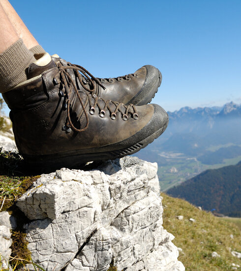 Das Bild zeigt die ledernen Bergschuhe einer Person, die auf einem Berg sitzt. Weiter unten erkennt man Bergwiesen, Wälder und ein Tal. Dahinter erheben sich weitere Berge. 