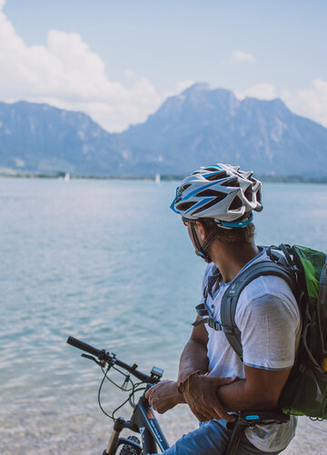 Das Bild fängt einen ruhigen Moment eines Radfahrers ein, der eine Pause von seiner Reise macht. Der Radfahrer lehnt auf seinem Fahrrad, das auf einem sandigen Strand geparkt ist. Der Radfahrer blickt auf den vor ihm liegenden Forggensee. Das Wasser ist ein tiefes Blau, das den klaren blauen Himmel darüber reflektiert. In der Ferne erheben sich majestätische Berge, die der Szene ein Gefühl von Größe verleihen. Der Radfahrer trägt einen Helm. Die Bild vermittelt eine ruhige Stimmung.