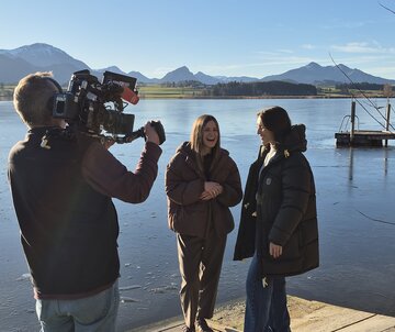 Es ist ein Kameramann sowie zwei Frauen, die ein Interview vor dem Panoram des Hopfensees führen, zu sehen.