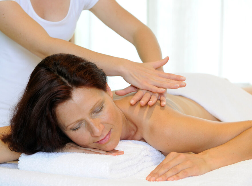 Das Bild zeigt eine Frau, die bäuchlings auf einer Massageliege liegt. Über ihr steht eine Person, die ihr die obere Rückenpartie massiert. 