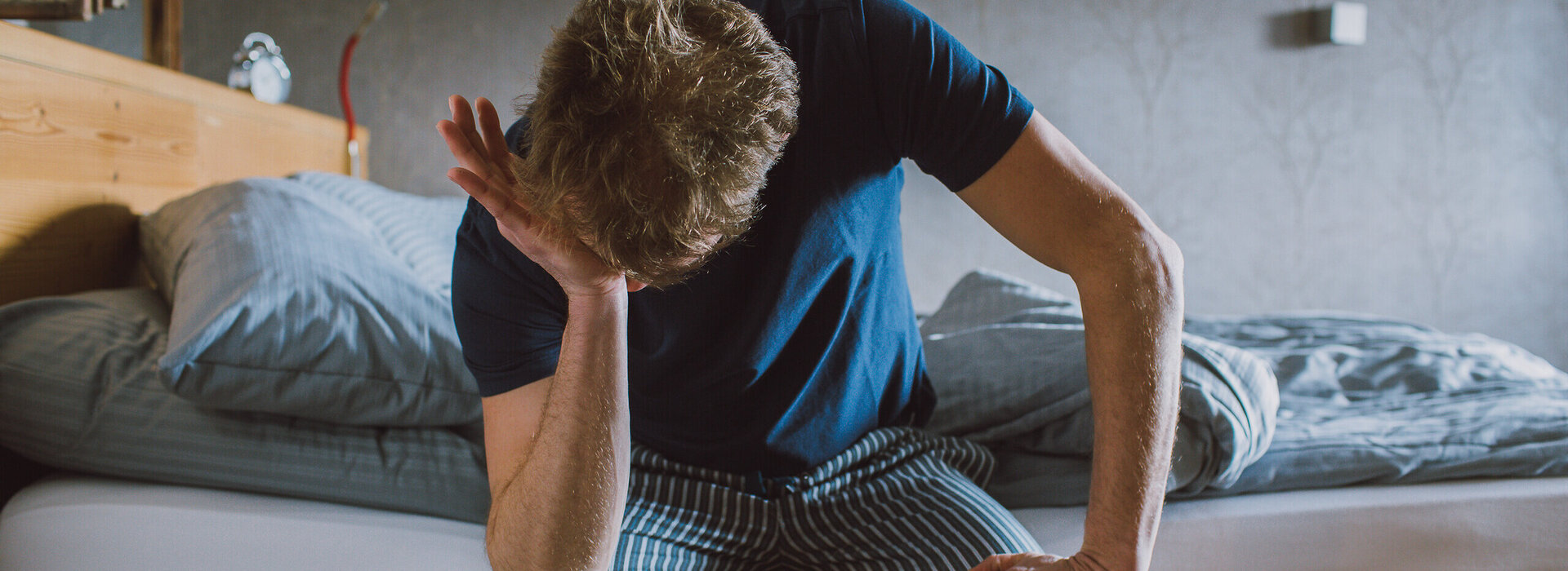 Das Bild zeigt einen Mann, der in einem Pyjama am Bettrand sitzt und die eine Hand auf den anderen Ellenbogen auf seine Knie gestützt hat. Mit der einen anderen Hand reibt er sich das Gesicht.