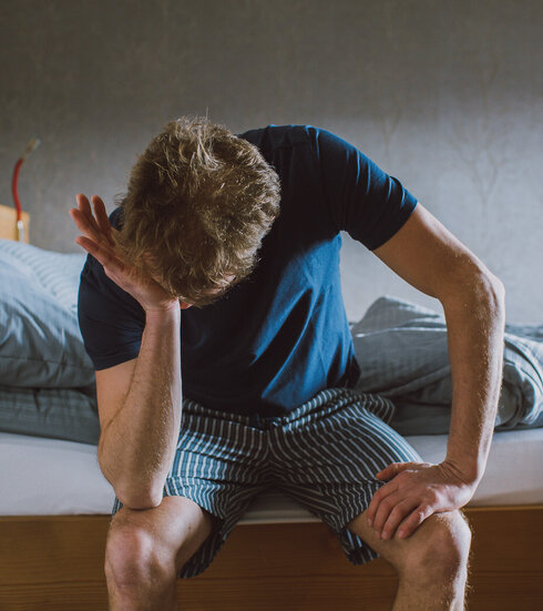 Das Bild zeigt einen Mann, der in einem Pyjama am Bettrand sitzt und die eine Hand auf den anderen Ellenbogen auf seine Knie gestützt hat. Mit der einen anderen Hand reibt er sich das Gesicht.
