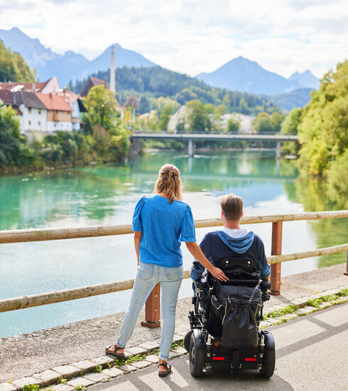 Das Bild fängt einen friedlichen Moment zweier Personen an einer Holzbrücke ein, die einen Blick auf eine Flusslandschaft bietet. Der Mann sitzt im Rollstuhl, während die Frau neben ihm steht. Im Hintergrund schmiegt sich ein malerisches Dorf in die üppige Vegetation. 