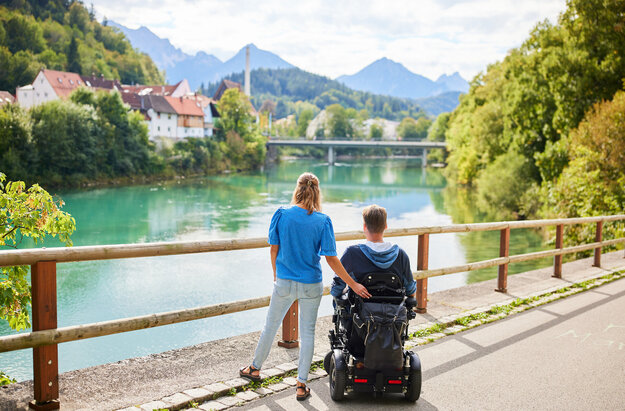 Das Bild fängt einen friedlichen Moment zweier Personen an einer Holzbrücke ein, die einen Blick auf eine Flusslandschaft bietet. Der Mann sitzt im Rollstuhl, während die Frau neben ihm steht. Im Hintergrund schmiegt sich ein malerisches Dorf in die üppige Vegetation. 
