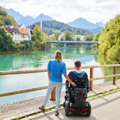 Das Bild fängt einen friedlichen Moment zweier Personen an einer Holzbrücke ein, die einen Blick auf eine Flusslandschaft bietet. Der Mann sitzt im Rollstuhl, während die Frau neben ihm steht. Im Hintergrund schmiegt sich ein malerisches Dorf in die üppige Vegetation. 