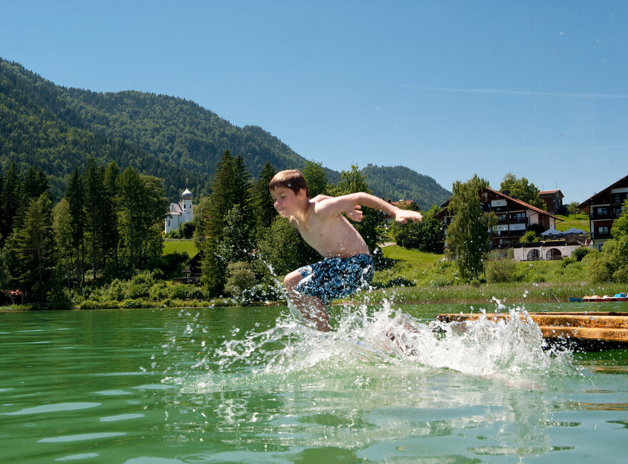 Das Bild zeigt einen Jungen, der in den Weißensee springt. Er hat die Wasseroberfläche bereits durchbrochen und das Wasser spritzt in alle Richtungen umher. Der Junge trägt blaue Badehosen. Der Weißensee ist von grünen Bäumen und Hügeln umgeben. Der Himmel ist blau. Die Sonne scheint.  