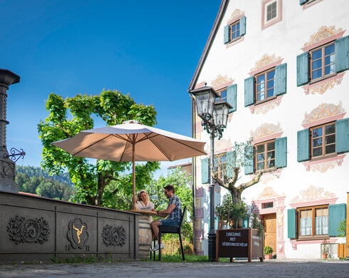 Ein Paar sitzt unter einem Sonnenschirm vor einem historischen Gebäude in der Füssener Altstadt. Die Szene wirkt idyllisch und ruhig.