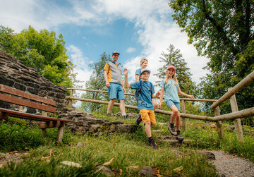 Das Bild zeigt eine Gruppe von vier Personen, zwei Jungen und zwei Mädchen, die auf einer hölzernen Treppe in einem Park stehen. Die Jungen sind mit blauen Hemden und khakifarbenen Shorts gekleidet, während die Mädchen blaue Oberteile und Denim-Shorts tragen. Alle haben ein Lächeln auf dem Gesicht und scheinen ihre gemeinsame Zeit zu genießen. Der Park ist grün und üppig, mit Bäumen und Rasen, die die Treppe umgeben. Der Himmel darüber ist ein klarer Blau, was auf einen sonnigen Tag hindeutet. Die hölzerne Treppe, die der Hauptfokus des Bildes ist, führt zu einer Steinmauer, die der Szene einen Hauch von Rustikalität verleiht. Die Atmosphäre des Bildes insgesamt ist eine von Freude und Kameradschaft, da die Gruppe im Park offensichtlich eine großartige Zeit miteinander hat.