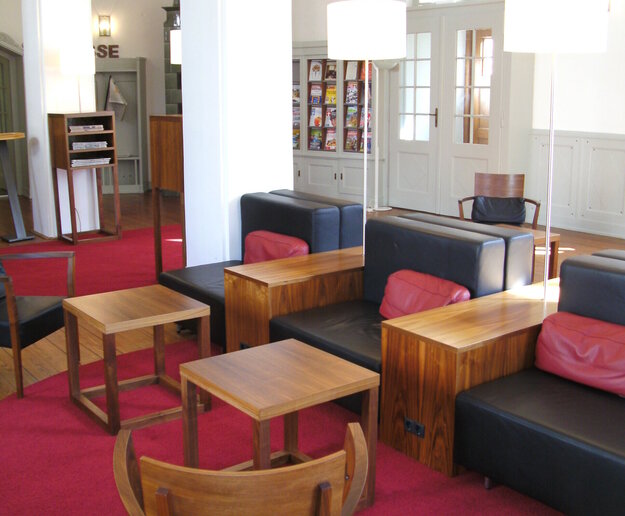 Das Bild zeigt einen Innenraum der Stadtbibliothek Füssen mit Holzmöbeln und roten Teppichboden. Eine Tür und Bücherregale sind im Hintergrund sichtbar.