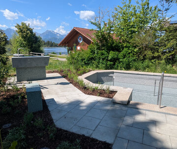 Ein neu gestalteter Außenbereich mit einem flachen Pool und einem Handlauf. Natursteinplatten bilden den Gehweg, umgeben von Büschen und kleinen Pflanzen. Im Hintergrund ist ein Wasserlauf sowie ein Holzgebäude sichtbar, das für Erholung genutzt werden kann. Die Landschaft ist von Bergen umgeben.