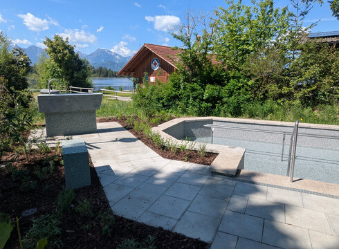 Kneipp-Anlage_Hopfen am See Ein neu gestalteter Außenbereich mit einem flachen Pool und einem Handlauf. Natursteinplatten bilden den Gehweg, umgeben von Büschen und kleinen Pflanzen. Im Hintergrund ist ein Wasserlauf sowie ein Holzgebäude sichtbar, das für Erholung genutzt werden kann. Die Landschaft ist von Bergen umgeben.