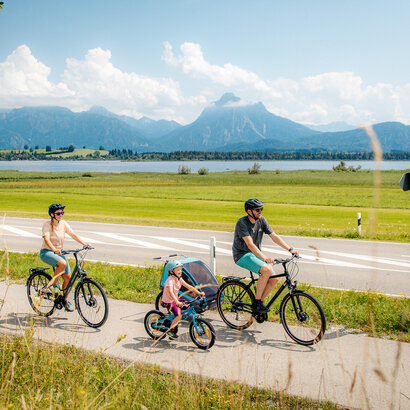 Das Bild fängt eine ruhige Szene von einer Familie mit einem Mann, einer Frau und einem kleinen Kind ein, die Fahrräder auf einem Pfad neben einer Straße fahren. Der Mann hat zudem noch einen Kinderanhänger am Fahrrad, in dem sich noch ein anderes Kleinkind befindet. Der Pfad ist von üppigem Grün umgeben. Im Hintergrund erkennt man den Hopfensee und die Berge. Der Himmel darüber ist ein klarer Blau mit fluffigen weißen Wolken. Die Radfahrer sind in legerer Kleidung und sommerlich angezogen. Sie tragen alle einen Helm. 