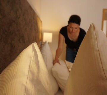 Das Bild zeigt eine Frau in Jeans und kurzem schwarzen T-Shirt, die ein Bett macht. Sie lehnt mit dem Oberkörper über dem Doppelbett und richtet die Kopfkissen. Die Bettdecken und Kissen sind weiß bezogen.  