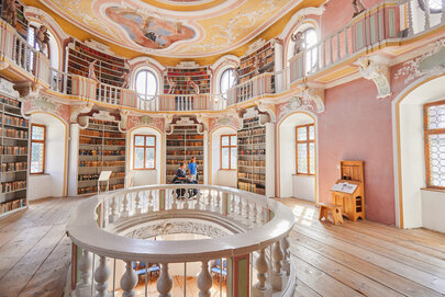 Die Bibliothek des Hohen Schlosses in Füssen mit hohen Regalen und einem beeindruckenden Rundbogen. Das Bild zeigt einen Raum voller Wissen und Geschichte.