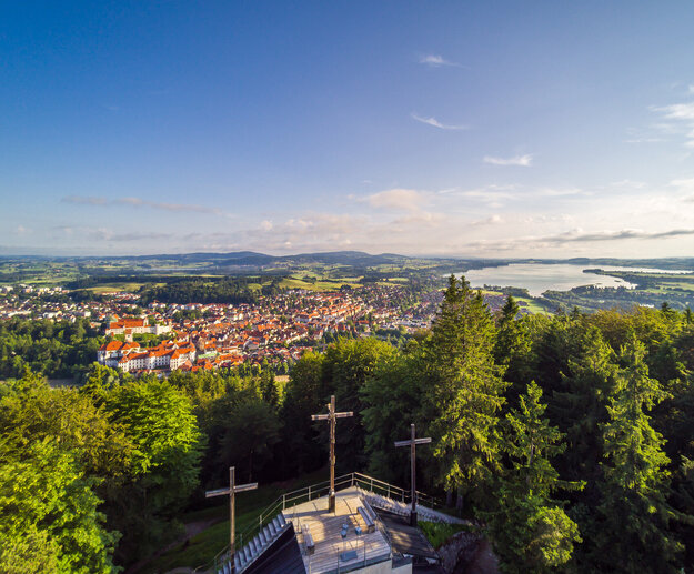 Das Bild zeigt einen Berg mit drei Kreuzen, die auf einer steinernen Aussichtsplattform stehen, aus der Vogelperspektive. Die Aussichtsplattform ist umgeben von hohen, grünen Bäumen. Am Fuß des Berges ist eine Stadt und Seen zu sehen.