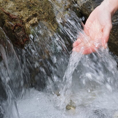Das Bild zeigt eine Nahaufnahme der Hand einer Person, die unter einem Wasserlauf gehalten wird. Das Wasser aus dem Bach fließt über einen felsigen Absatz in einem kleinen Wasserfall sprudelnd nach unten. 