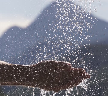 Das Bild zeigt zwei zusammengehaltene Hände in die Wasser einfließt. Im Hintergrund erkennt man einen markannten Berg.