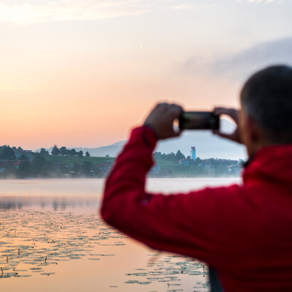 A man stands in front of a lake in the sunset and photographs the atmosphere with his cell phone. There is a light mist over the water