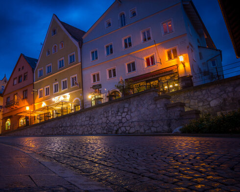 Das Bild zeigt ein Gebäude in der Füssener Altstadt mit vielen beleuchteten Fenstern. Der Boden spiegelt das Licht wider.