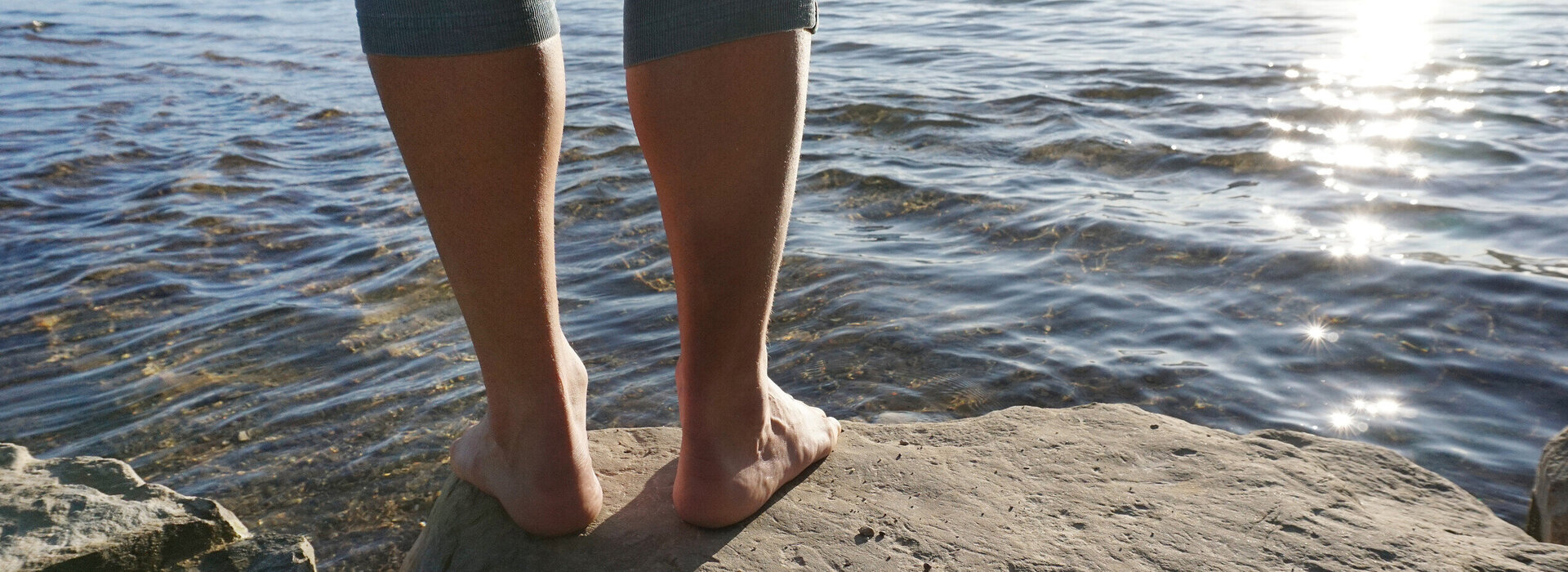 Das Bild zeigt einen Menschen, der barfuß auf einem Felsen vor dem Weißensee steht. Es sind lediglich seine Beine zu sehen. Im Wasser kräuseln sich kleine Wellen und die hell aufgehende Sonne glitzert darin.