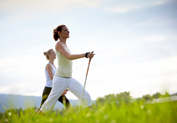 Das Bild zeigt zwei sportlich gekleidete, schlanke Frauen, die mit Nordic-Walking-Stöcken über eine grüne Wiese laufen. Der blaue Himmel ist von sanften Wolken durchzogen.