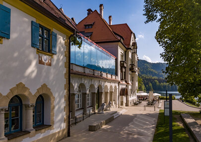 Das Museum der bayerischen Könige am Alpseeufer mit belebter Promenade und Blick auf die Berge.