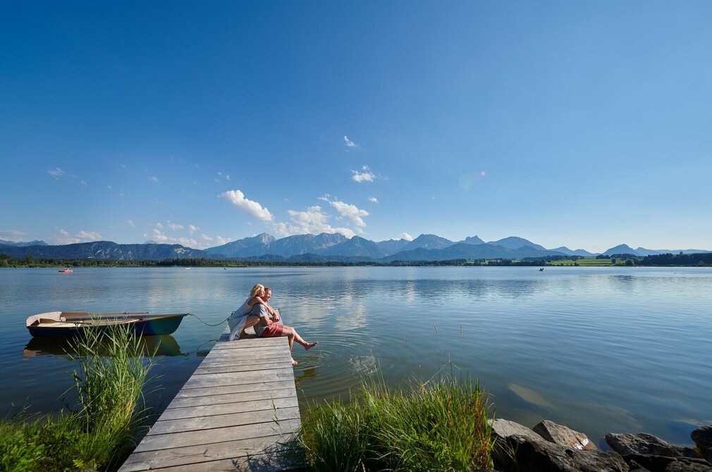 Auf dem Bild ist eine idyllische Szene am Hopfensee zu sehen. Eine Frau kniet hinter einem Mann auf einem Holzsteg und umarmt ihn von hinten. In der Ferne sind ein paar Ruder- und Tretboote auf dem Wasser zu sehen.