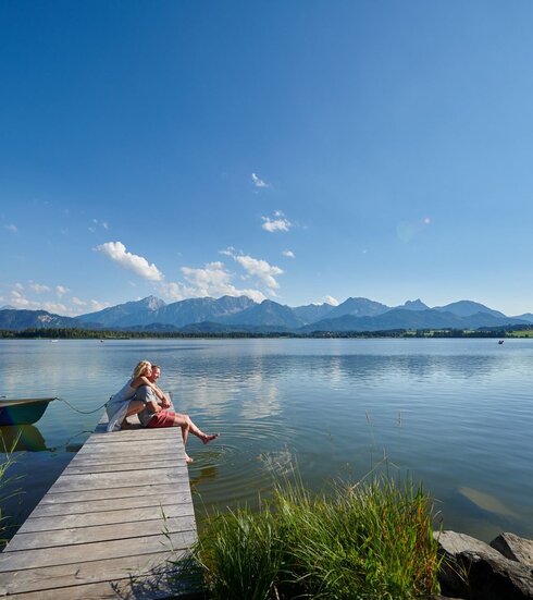 Auf dem Bild ist eine idyllische Szene am Hopfensee zu sehen. Eine Frau kniet hinter einem Mann auf einem Holzsteg und umarmt ihn von hinten. In der Ferne sind ein paar Ruder- und Tretboote auf dem Wasser zu sehen.