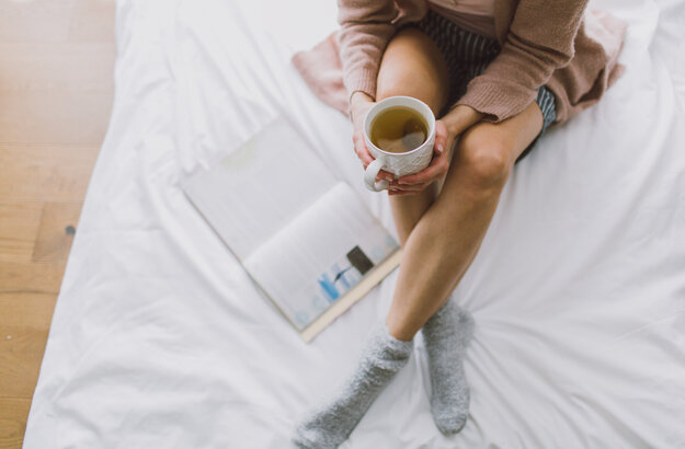 Das Bild zeigt eine Frau, die mit gekreuzten Beinen auf einem weiß bezogenen Bett sitzt. Sie hält eine Teetasse in der Hand. Neben ihr liegt ein Buch im Bett. 