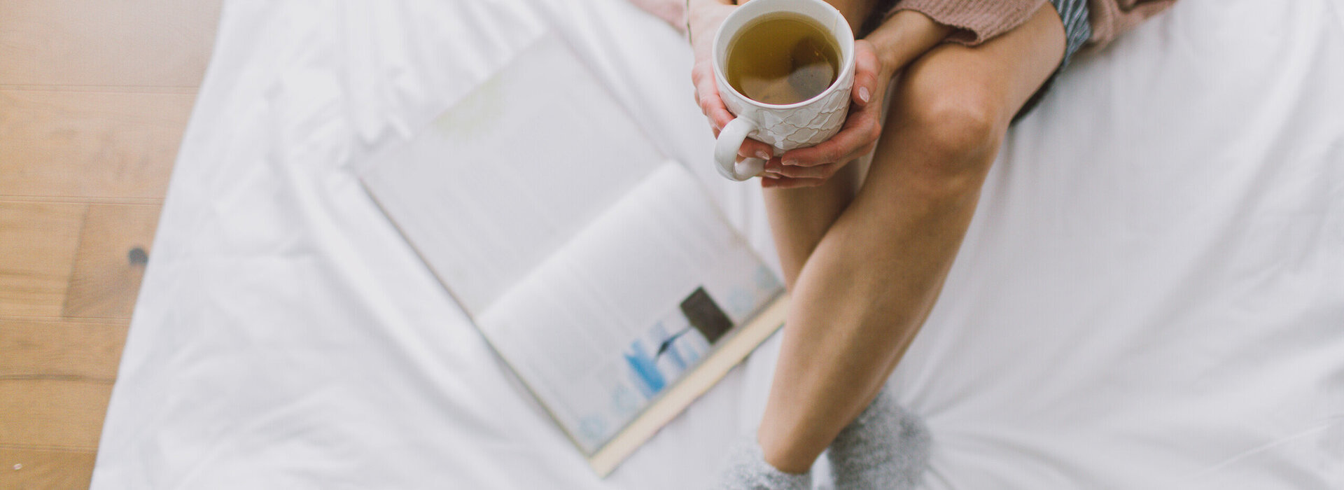 Das Bild zeigt eine Frau, die mit gekreuzten Beinen auf einem weiß bezogenen Bett sitzt. Sie hält eine Teetasse in der Hand. Neben ihr liegt ein Buch im Bett. 