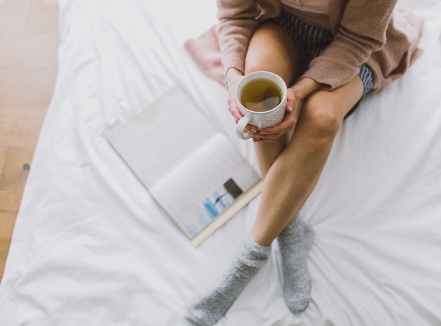 Das Bild zeigt eine Frau, die mit gekreuzten Beinen auf einem weiß bezogenen Bett sitzt. Sie hält eine Teetasse in der Hand. Neben ihr liegt ein Buch im Bett. 