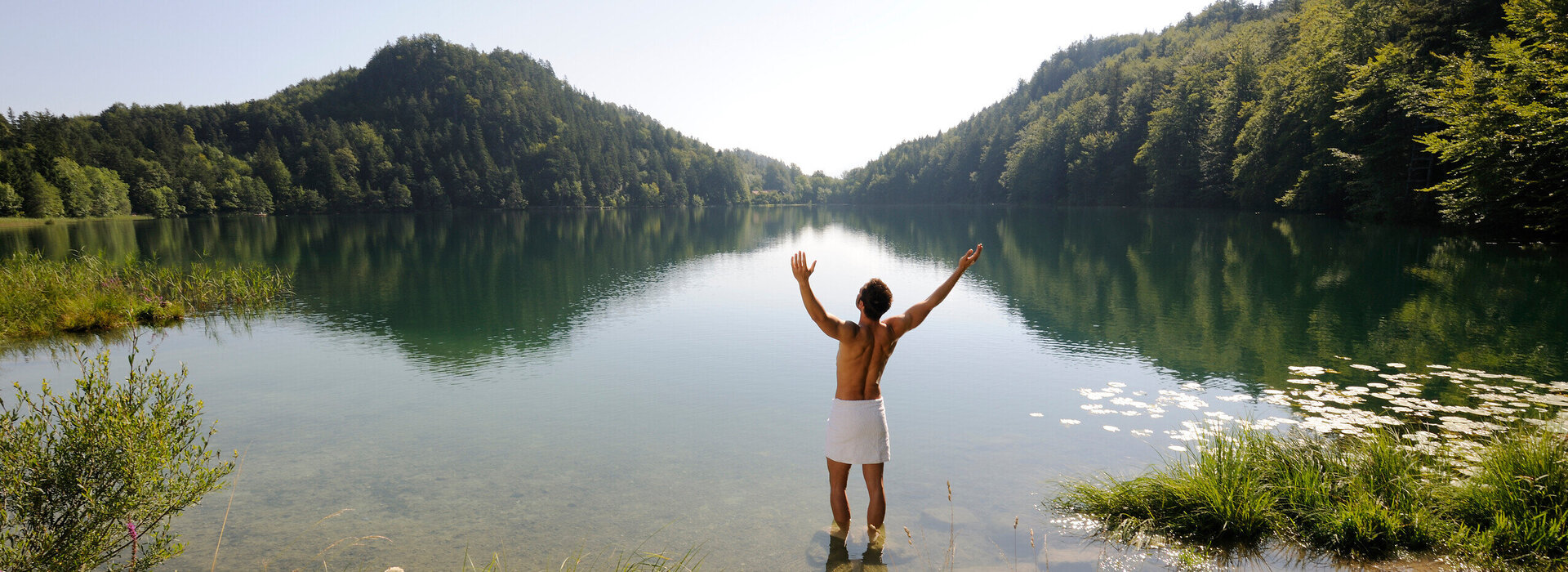 Das Bild zeigt einen Mann, der sich nur ein weißes Handtuch um die Hüften gebunden hat. Er steht im knöcheltiefen Wasser eines Sees und streckt die Arme nach oben.