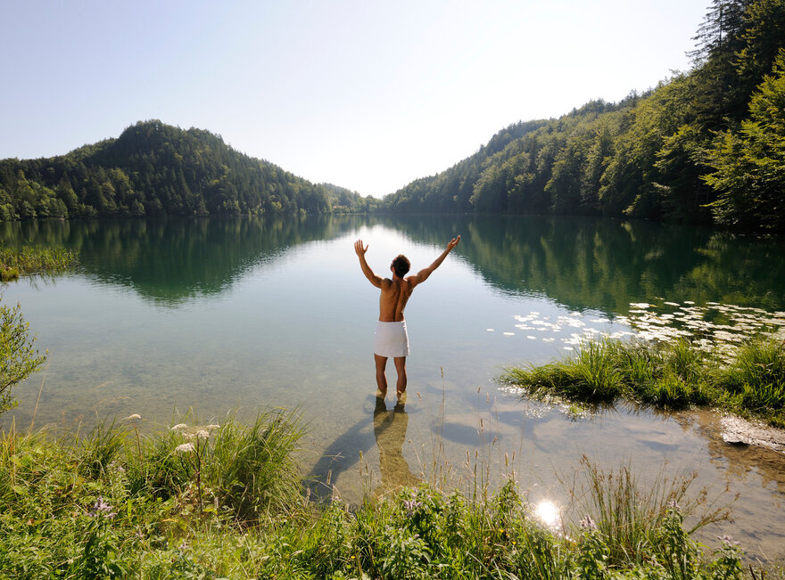 Das Bild zeigt einen Mann, der sich nur ein weißes Handtuch um die Hüften gebunden hat. Er steht im knöcheltiefen Wasser eines Sees und streckt die Arme nach oben.