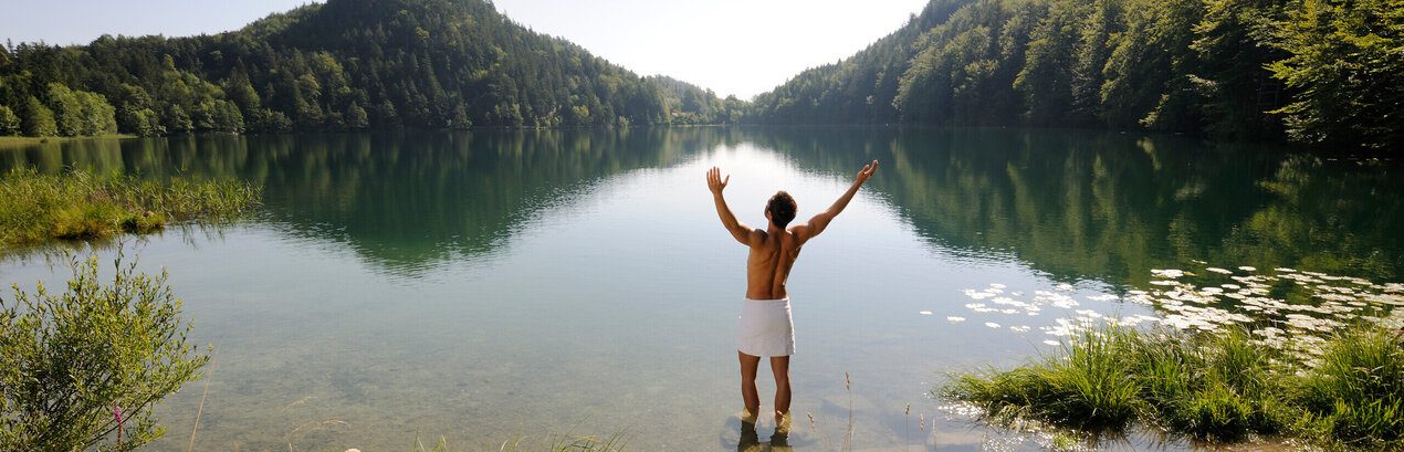 Das Bild zeigt einen Mann, der sich nur ein weißes Handtuch um die Hüften gebunden hat. Er steht im knöcheltiefen Wasser eines Sees und streckt die Arme nach oben.