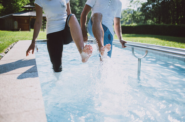 Das Bild zeigt zwei Personen, die in einem Kneipptretbecken stehen. Beide Personen heben ein Bein aus dem Wasser, sodass die Wassertropfen um sie herum spritzen. 