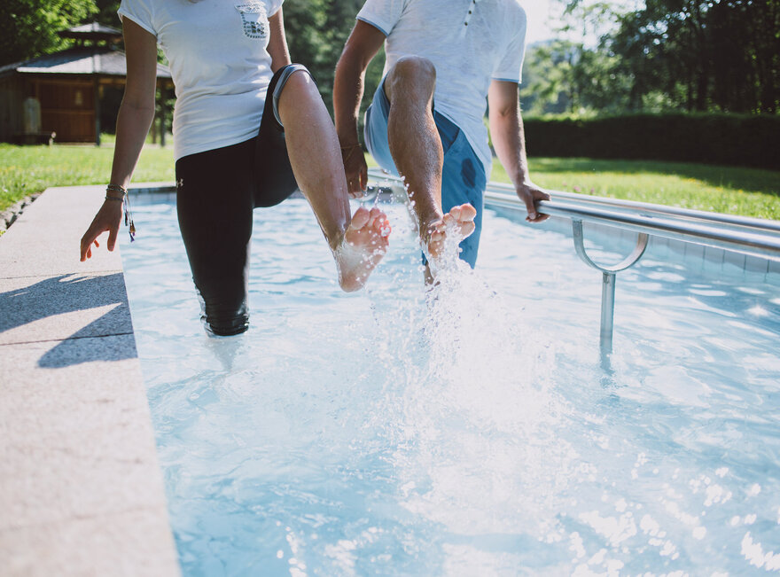 Das Bild zeigt zwei Personen, die in einem Kneipptretbecken stehen. Beide Personen heben ein Bein aus dem Wasser, sodass die Wassertropfen um sie herum spritzen. 
