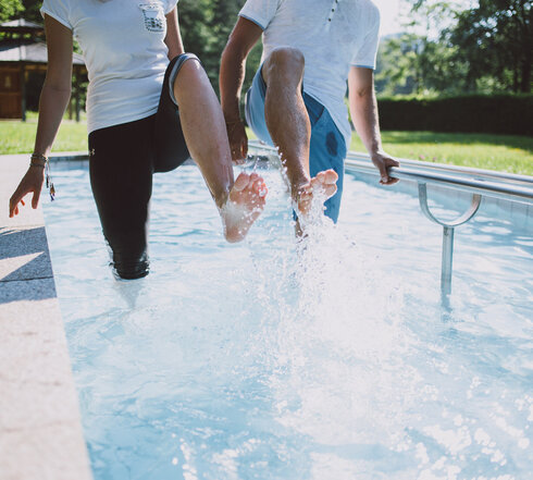Das Bild zeigt zwei Personen, die in einem Kneipptretbecken stehen. Beide Personen heben ein Bein aus dem Wasser, sodass die Wassertropfen um sie herum spritzen. 