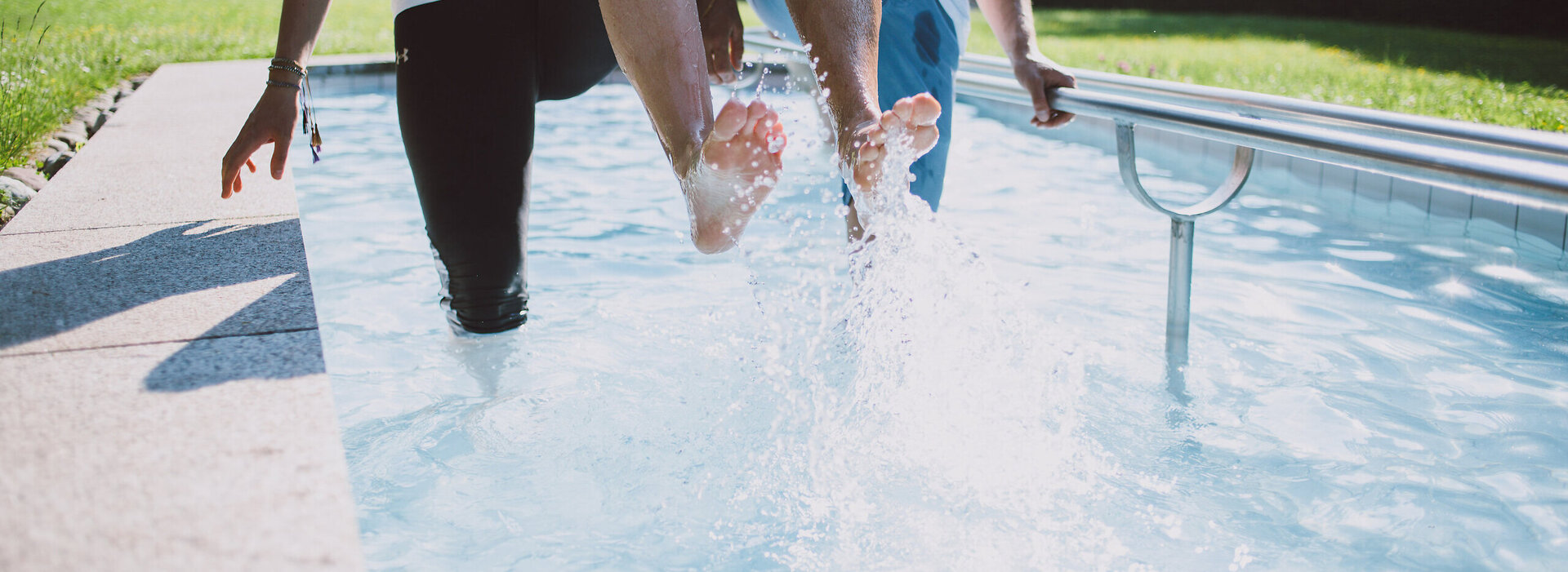 Das Bild zeigt zwei Personen, die in einem Kneipptretbecken stehen. Beide Personen heben ein Bein aus dem Wasser, sodass die Wassertropfen um sie herum spritzen. 