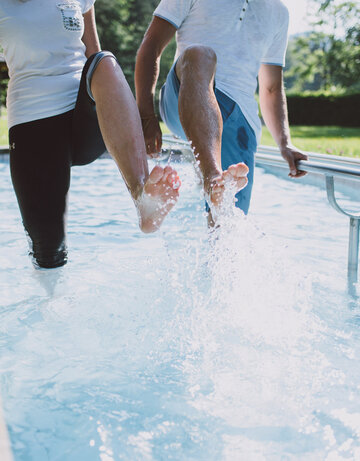 Das Bild zeigt zwei Personen, die in einem Kneipptretbecken stehen. Beide Personen heben ein Bein aus dem Wasser, sodass die Wassertropfen um sie herum spritzen. 