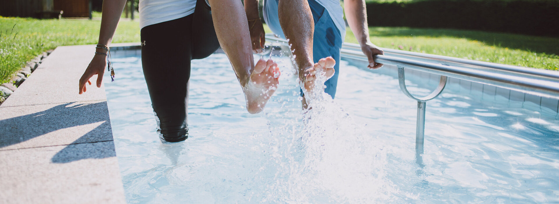 Das Bild zeigt zwei Personen, die in einem Kneipptretbecken stehen. Beide Personen heben ein Bein aus dem Wasser, sodass die Wassertropfen um sie herum spritzen. 