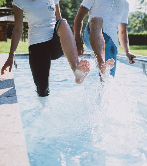 Das Bild zeigt zwei Personen, die in einem Kneipptretbecken stehen. Beide Personen heben ein Bein aus dem Wasser, sodass die Wassertropfen um sie herum spritzen. 