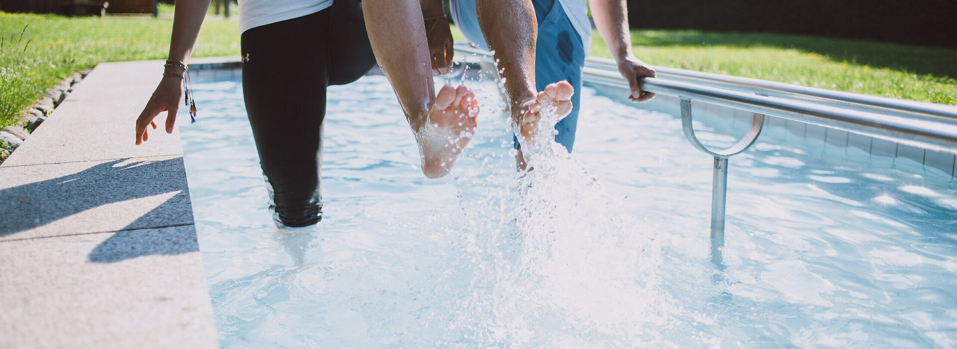 Das Bild zeigt zwei Personen, die in einem Kneipptretbecken stehen. Beide Personen heben ein Bein aus dem Wasser, sodass die Wassertropfen um sie herum spritzen. 