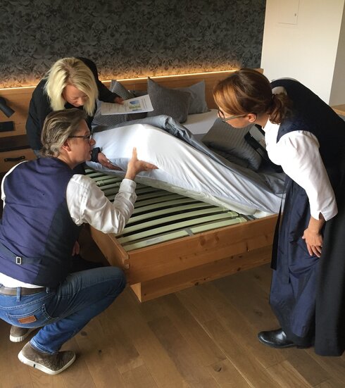 Das Bild zeigt drei Personen, die die Matratze eines Bettes hochheben und den darunterliegenden Lattenrost betrachten und überprüfen. 