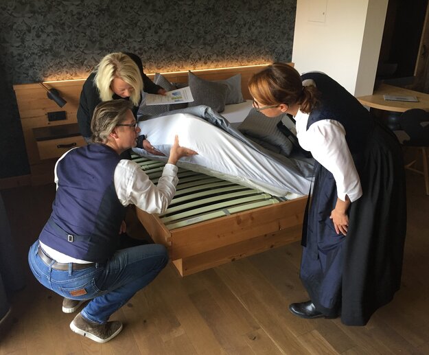 Das Bild zeigt drei Personen, die die Matratze eines Bettes hochheben und den darunterliegenden Lattenrost betrachten und überprüfen. 