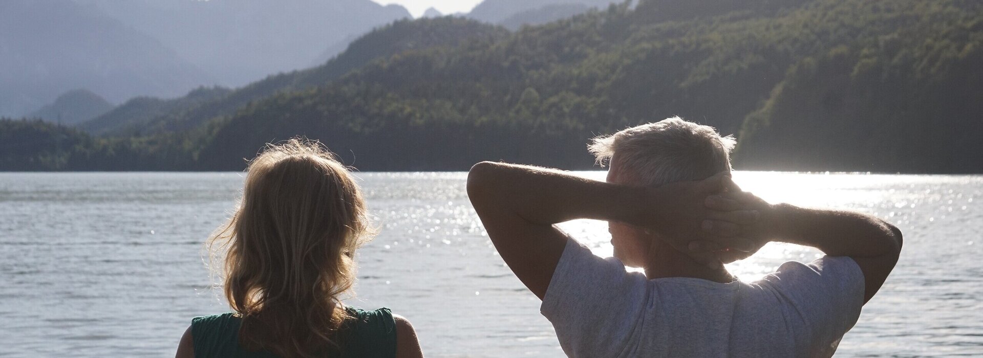 Das Bild zeigt einen Mann und eine Frau, die entspannt auf einer Bank sitzen und in die Ferne blicken. Im Hintergrund glitzert der Weißensee im Sonnenlicht und in der Ferne erhebt sich die markante Bergsilhouette des Säulings.